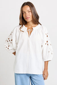 Model wearing the Betty Basics Daisy Broderie Blouse in white with floral cut-out puff sleeves, styled with light blue wide-leg jeans.