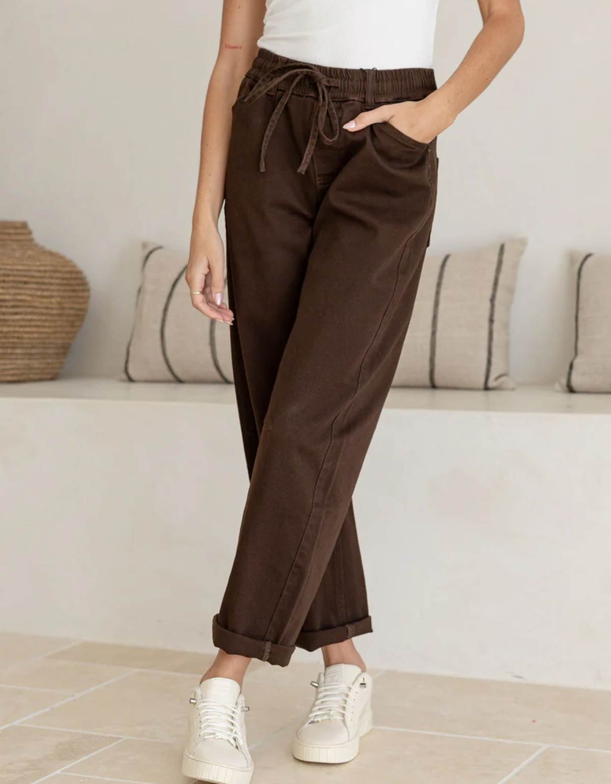 Country Denim Drawstring Barrel Leg Denim Jogger in Chocolate – cropped waist-to-ankle front detail view showing elasticated ruched waistband with self-fabric drawstring bow, side slash pocket, front panel seams with contrast topstitching, barrel leg silhouette and rolled cuff hem in chocolate brown stretch denim, with white platform sneakers on travertine tile