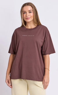 A woman modeling an oversized short-sleeve t-shirt in a washed dark brownish-purple color, featuring the text 'FOXWORD ATHLETICS' printed in white across the chest. She is wearing light beige pants against a plain light gray studio background.
