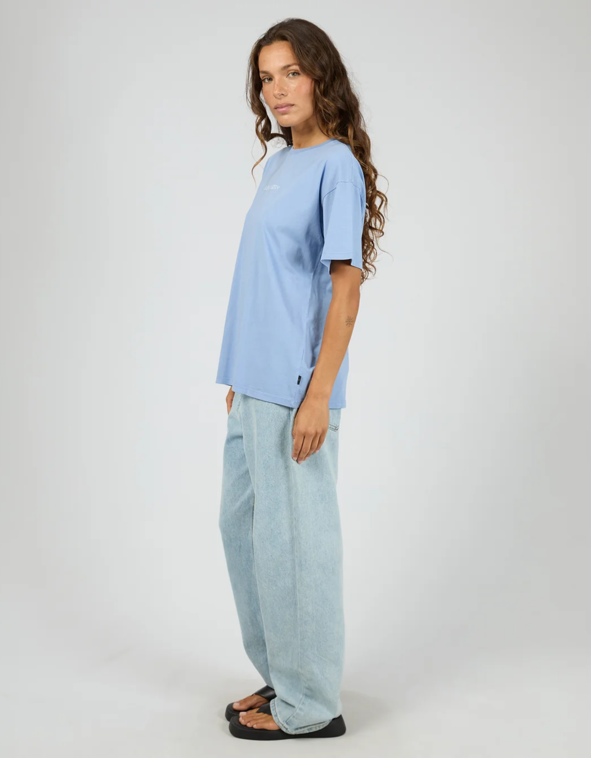 Silent Theory Logo Tee in Light Blue – full-length side profile showing relaxed boxy oversized silhouette, small side hem badge detail, styled with wide-leg light wash jeans and black thong sandals