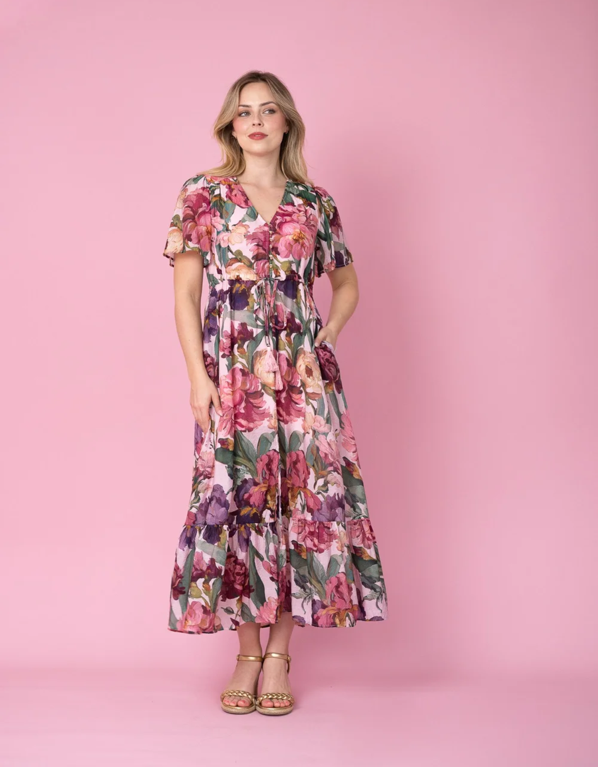 Model wearing the Fria The Label Selma Maxi Dress in Pink Floral, showing flutter sleeves, a tie waist and tiered skirt.